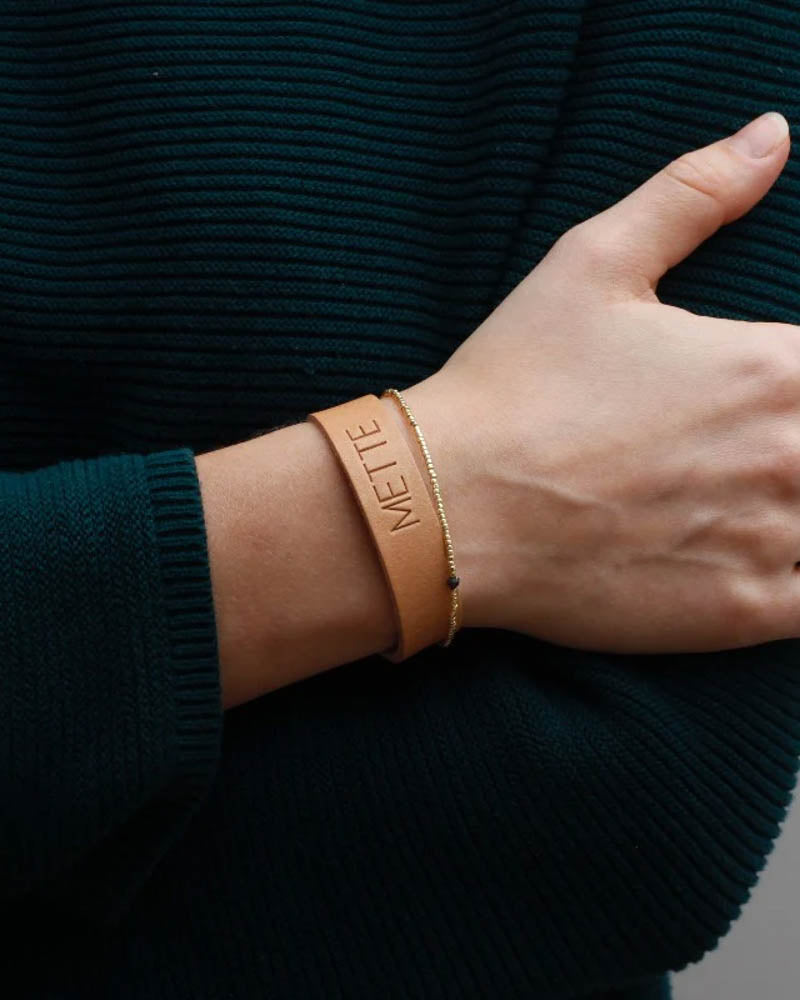 MONO läderarmband i naturfärgat läder med namnet "Mette" graverat runt handleden tillsammans med tunt guldarmband