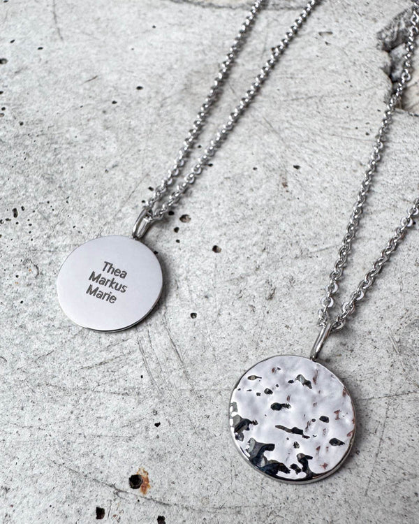 MOON QUOTE namnhalsband i silverfärgat rostfritt stål med namnen "Thea Markus Marie" graverade, hamrad sida visad bredvid, på betong