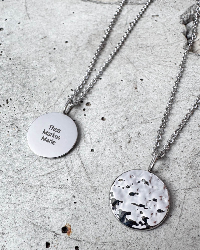 MOON QUOTE namnhalsband i silverfärgat rostfritt stål med namnen "Thea Markus Marie" graverade, hamrad sida visad bredvid, på betong