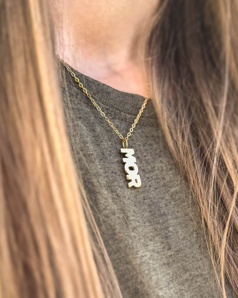 MOM halsband i guldpläterat stål med bokstavshänget "MOR", buret runt halsen över grå t-shirt