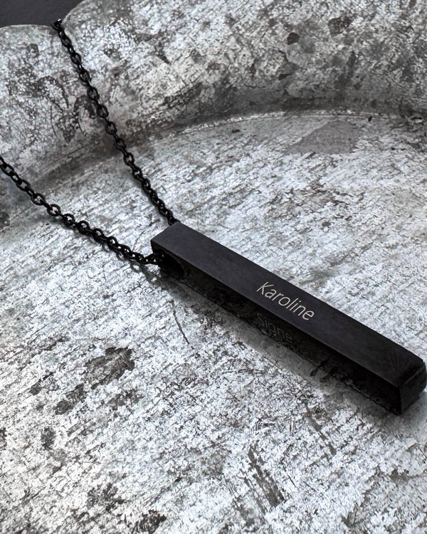 NEO namnhalsband i svart stål med namnet "Karoline" graverat på avlång stav, liggande på patinerat zinkfat