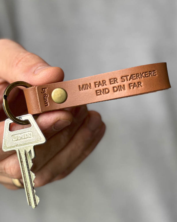 RAVN nyckelring i cognac läder med gravyren "Min far er stærkere end din far", hålls i handen med nyckel