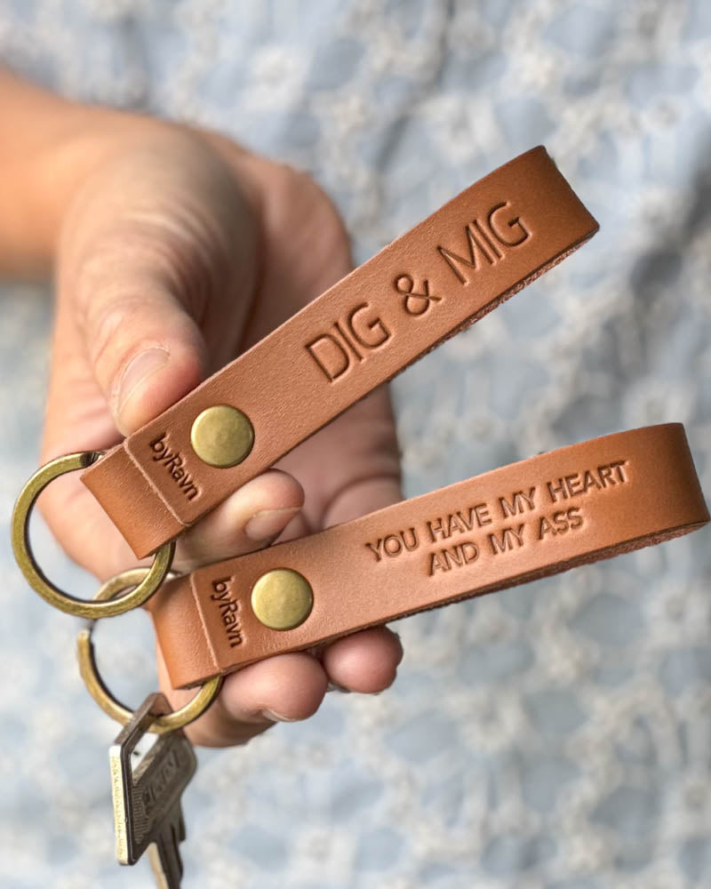 Två RAVN nyckelringar i cognac läder som hålls i handen med gravyrerna "Dig & mig" och "You have my heart and my ass"