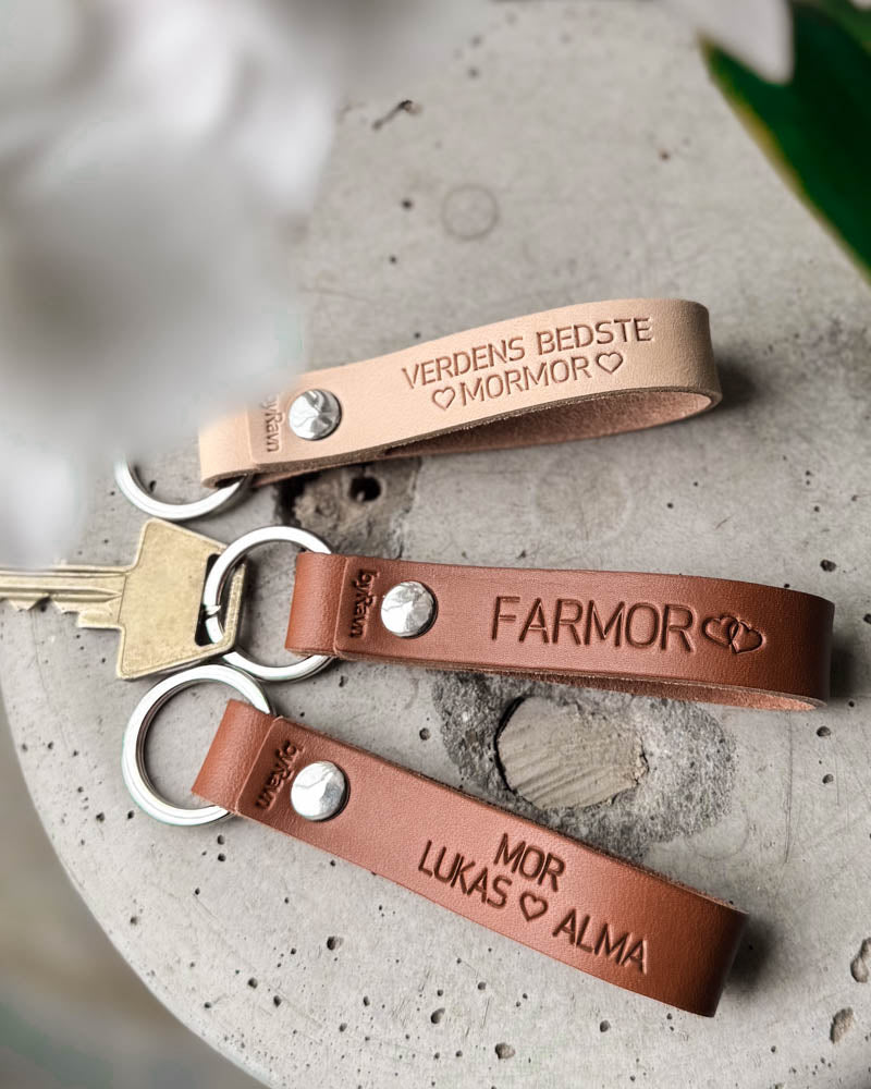 Tre RAVN nyckelringar i naturfärgat och cognac läder med "Verdens bedste mormor", "Farmor" och "Mor Lukas ♡ Alma" graverat, på betong