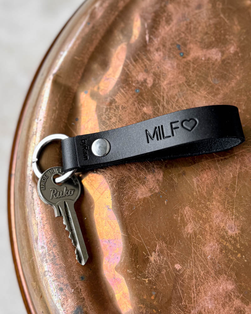RAVN nyckelring i mörkt läder med gravyren "MILF" ♡ och nyckel, liggande på patinerad kopparbricka