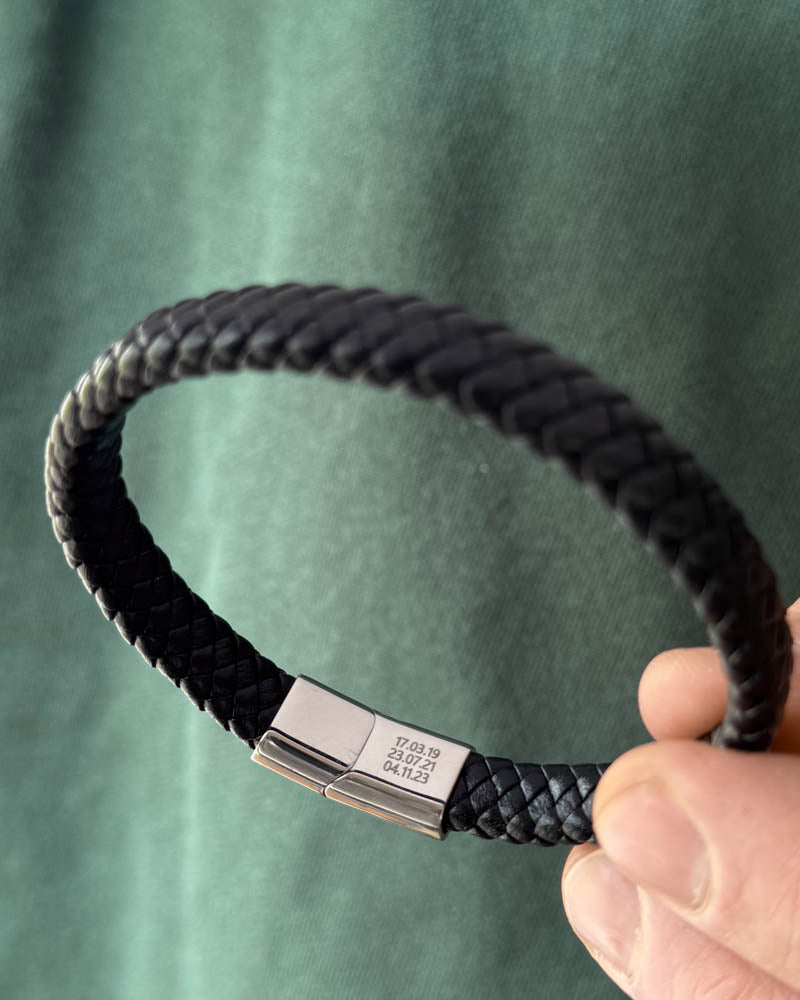 RAY läderarmband i svart flätat läder som hålls i handen, datumen "17.03.19, 23.07.21, 04.11.23" graverade på låset
