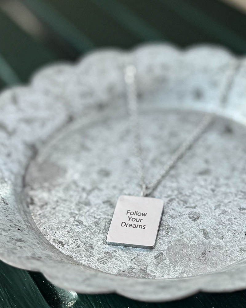 RAZ halsband i silverfärgat rostfritt stål med gravyren "Follow Your Dreams" på rektangulär bricka, på vitpatinerat zinkfat