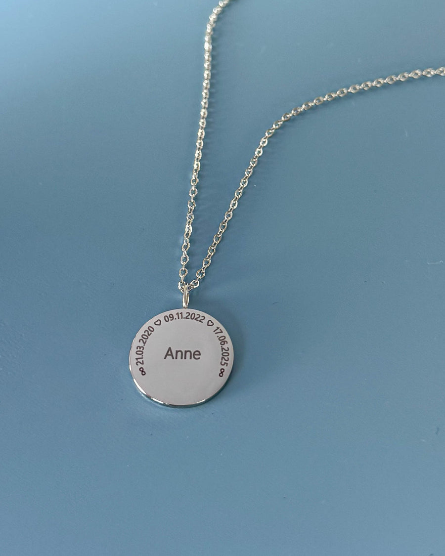 SIA CIRCLE namnhalsband i silverfärgat rostfritt stål med "Anne" och datumen "21.03.2020 ♡ 09.11.2022 ♡ 17.06.2025" i cirkel, på blå botten