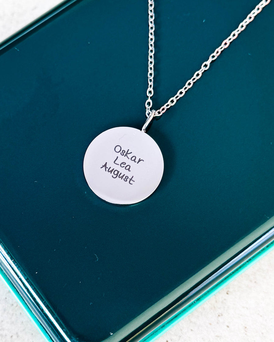 SIA QUOTE SCRIPT namnhalsband i silverfärgat rostfritt stål med namnen "Oskar Lea August" i handskrift, på grön glasask
