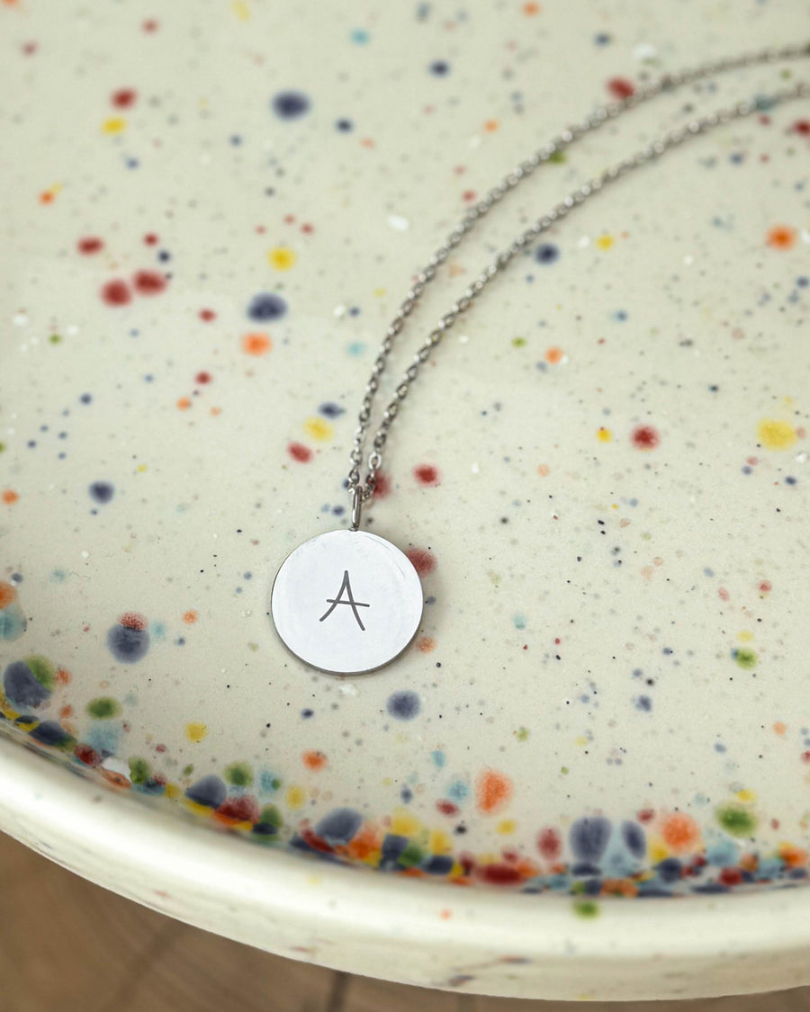 SIA LETTER SCRIPT halsband i silverfärgat rostfritt stål med bokstaven "A" i handskrift graverad, på prickigt keramikfat