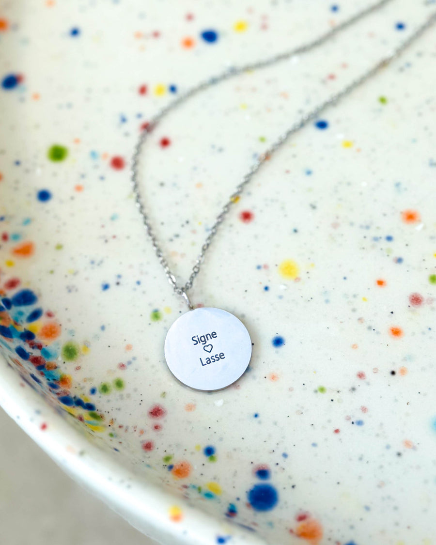 SIA QUOTE namnhalsband i silverfärgat rostfritt stål med gravyren "Signe ♡ Lasse" på runt hänge, på prickigt fat