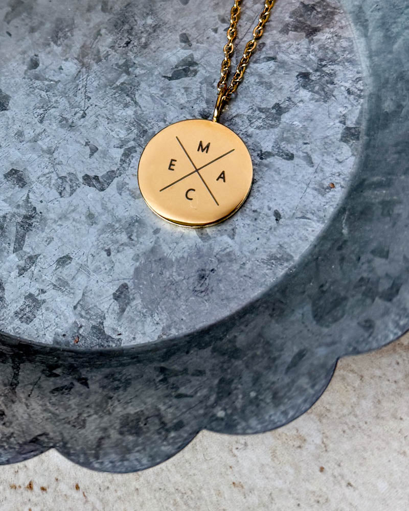 SIA X halsband i guldpläterat stål med initialerna "M E A C" graverade i kors på runt hänge, liggande på zinkfat