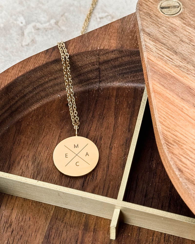 SIA X halsband i guldpläterat stål med bokstäverna "M E A C" i kors på runt hänge, i träskål med mässingskant