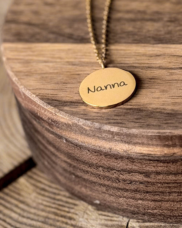 SIA NOTE SCRIPT namnhalsband i guldpläterat stål med namnet "Nanna" i handskrift graverat på runt hänge, på runt träfat