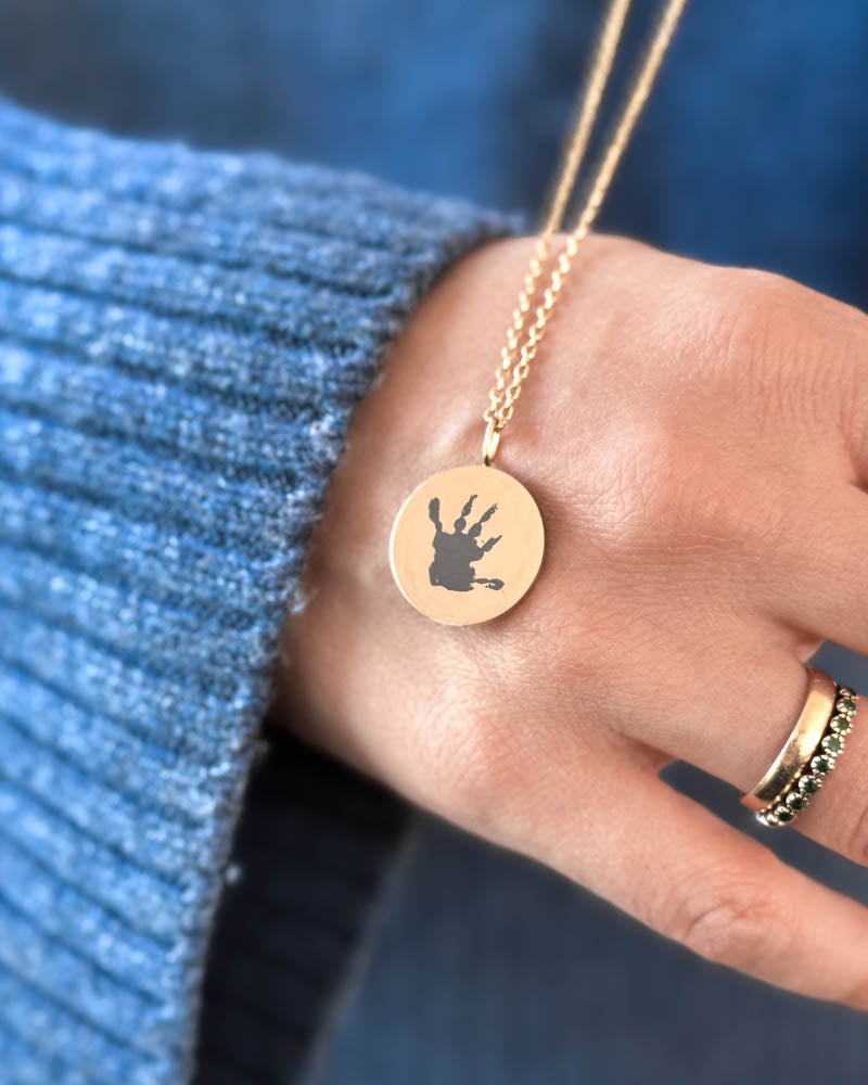 SIA MOTIF halsband i guldpläterat stål med ett litet handavtryck graverat på runt hänge, hålls i handen över blå stickat