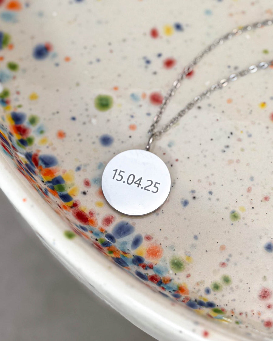 SIA NOTE halsband i silverfärgat rostfritt stål med datumet "15.04.25" graverat på runt hänge, på prickigt keramikfat
