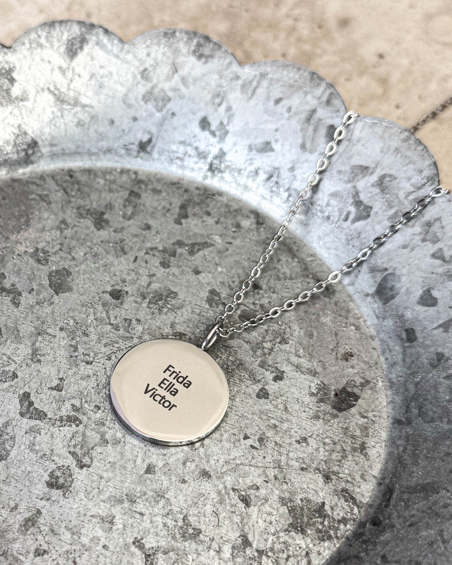 SIA QUOTE namnhalsband i silverfärgat rostfritt stål med namnen "Frida, Ella, Victor" graverade, liggande på zinkfat