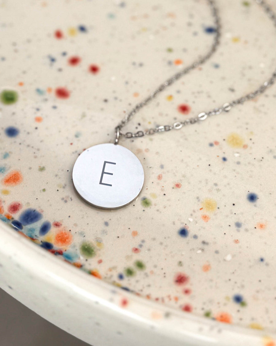 SIA LETTER halsband i silverfärgat rostfritt stål med bokstaven "E" graverad på runt hänge, på prickigt keramikfat