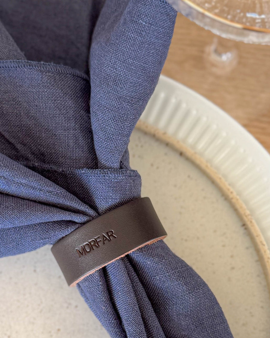 KNOT servettring placeringskort i mörkbrunt läder med gravyren "Morfar" runt mörkblå linneservett på räfflad tallrik
