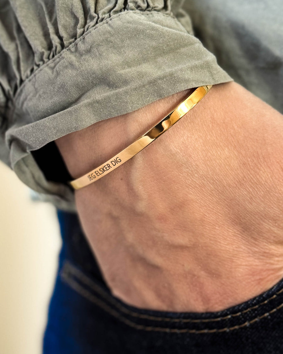 VENUS armband i guldpläterat stål med gravyren "Jeg elsker dig" runt handleden, hand i fickan på mörka jeans