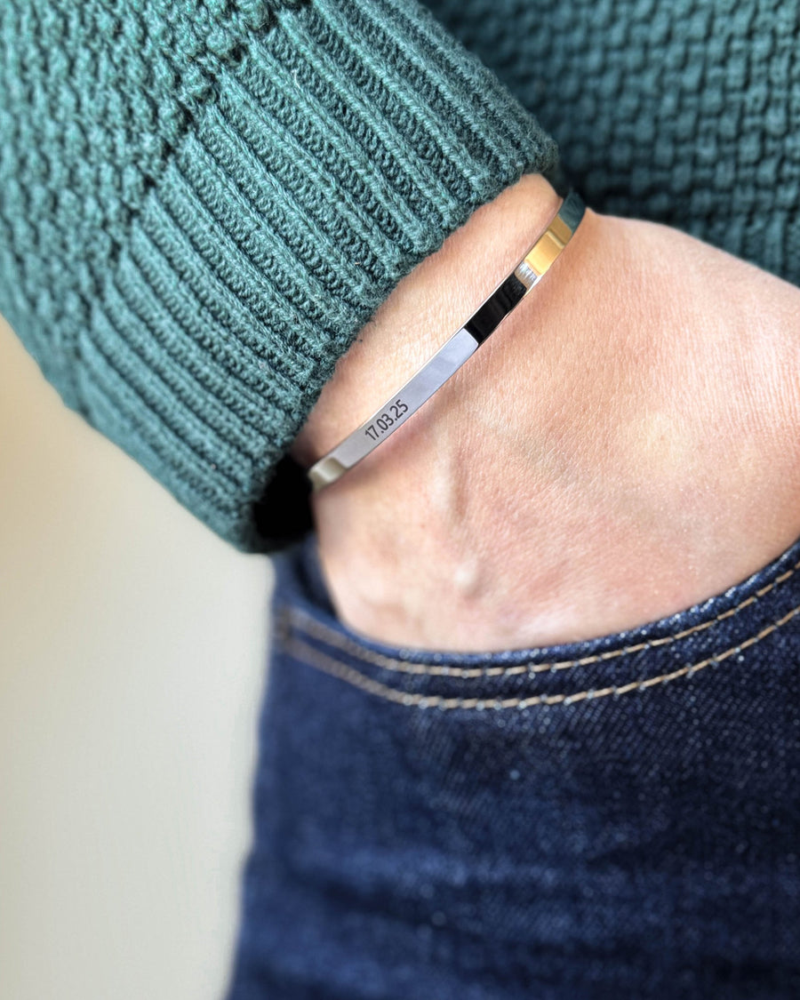 VENUS armband i silverfärgat rostfritt stål med datumet "17.03.25" graverat, hand i fickan på mörka jeans, grön stickad tröja