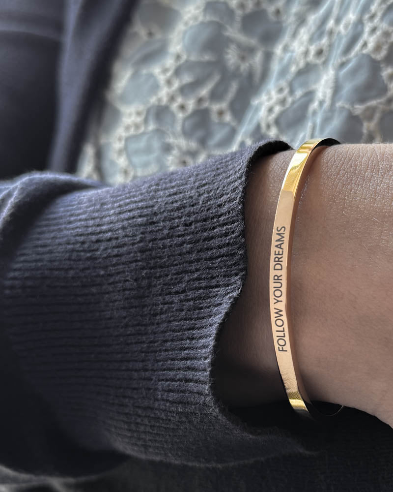 VENUS armband i guldpläterat stål med gravyren "Follow your dreams" i versaler, buret till mörkblå stickat och spetsblus