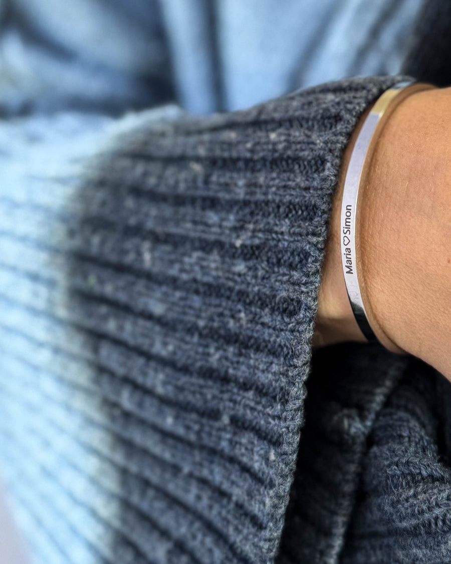 VENUS armband i silverfärgat rostfritt stål med gravyren "Maria ♡ Simon" runt handleden, blå stickad tröja